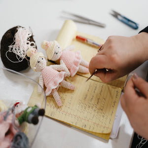 artisan hands holding knitting needle reviewing notepad with numbers. small knitted dolls lay over notepad.